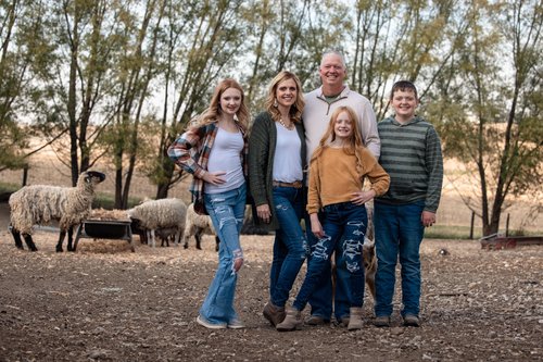 Family portrait on a farm with sheep in the background, featuring two adults and three children dressed in casual attire, surrounded by trees and rustic scenery.
