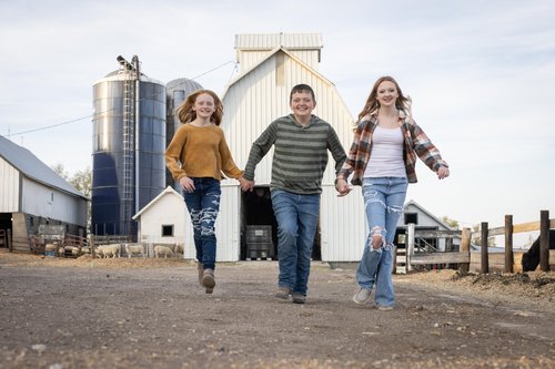 Three children running joyfully on a farm path, holding hands, with a barn and silos in the background. The scene captures a playful moment in a rural setting, showcasing the beauty of farm life.