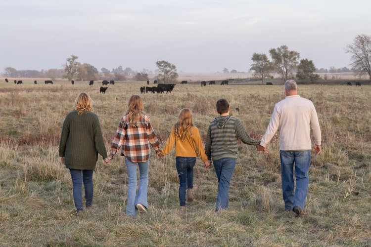 Family walking hand-in-hand through a rural field with cows in the background, showcasing a serene outdoor lifestyle. The group includes two adults and three children, dressed in cozy, casual attire, enjoying a peaceful moment in nature.